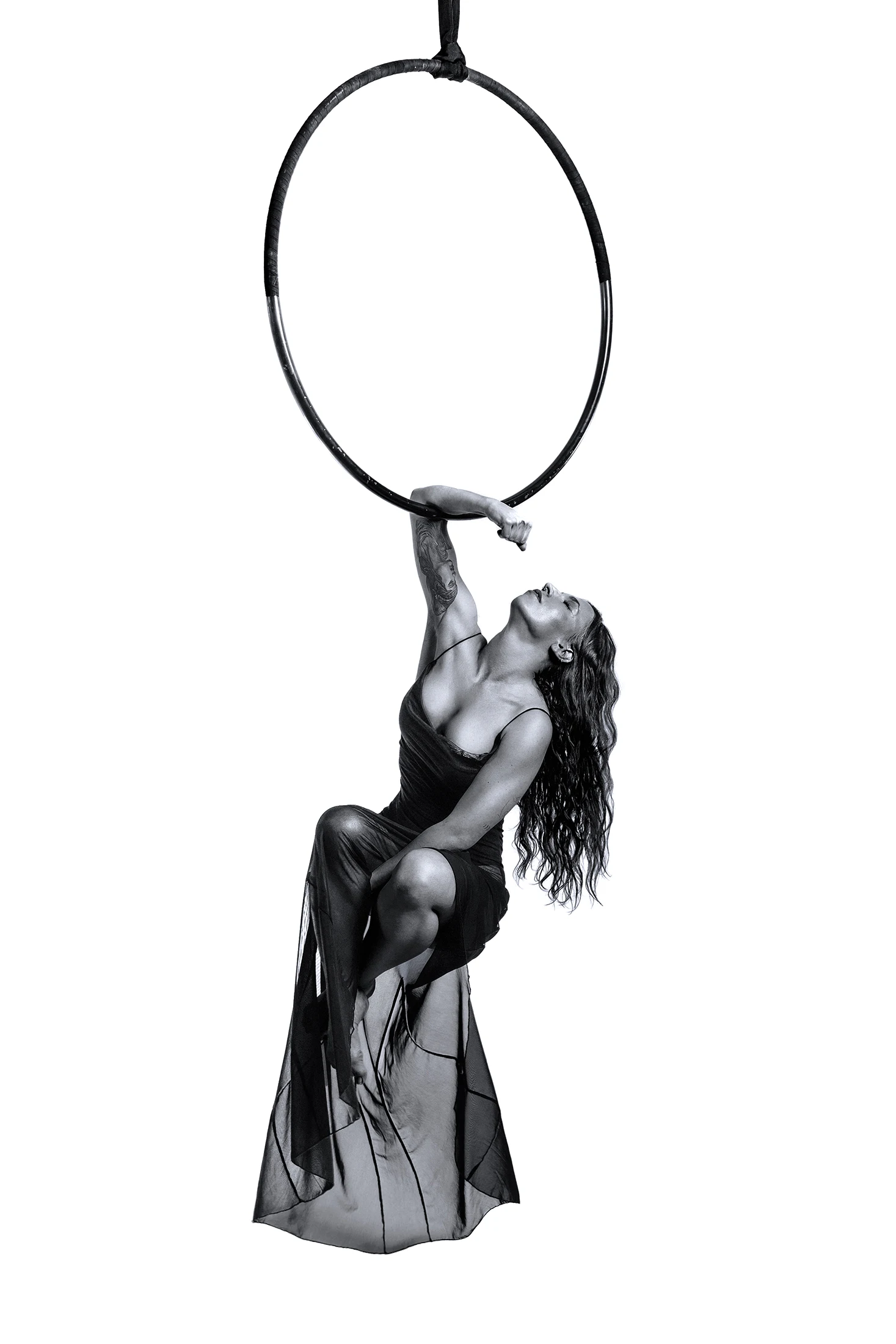 Aerialist in a long sheer dress hanging by one arm beneath an empty hoop.