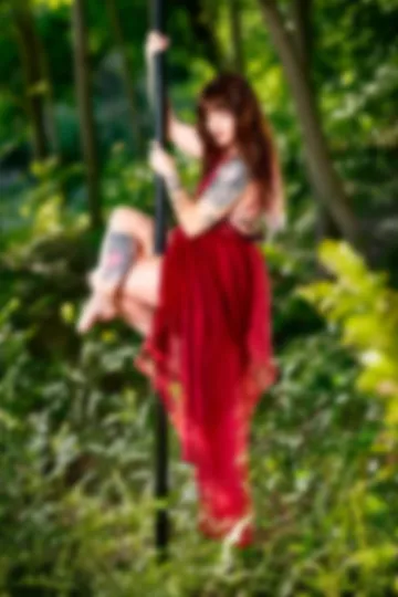 Pole dancer in a red tulle dress seated on the pole with legs crossed, in a green forest.