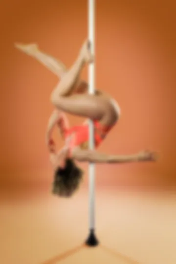 Inverted on the pole with legs folded and arms spread wide, in an orange bodysuit against an orange ground.