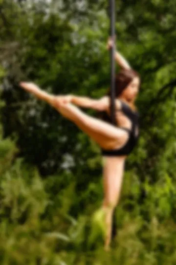 Dancer in black beside the pole, one leg stretched out horizontally and held in her hand, among forest foliage.