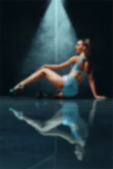 Seated on a mirror-dark floor beside the pole, legs long in platform heels, under a single shaft of icy blue haze.