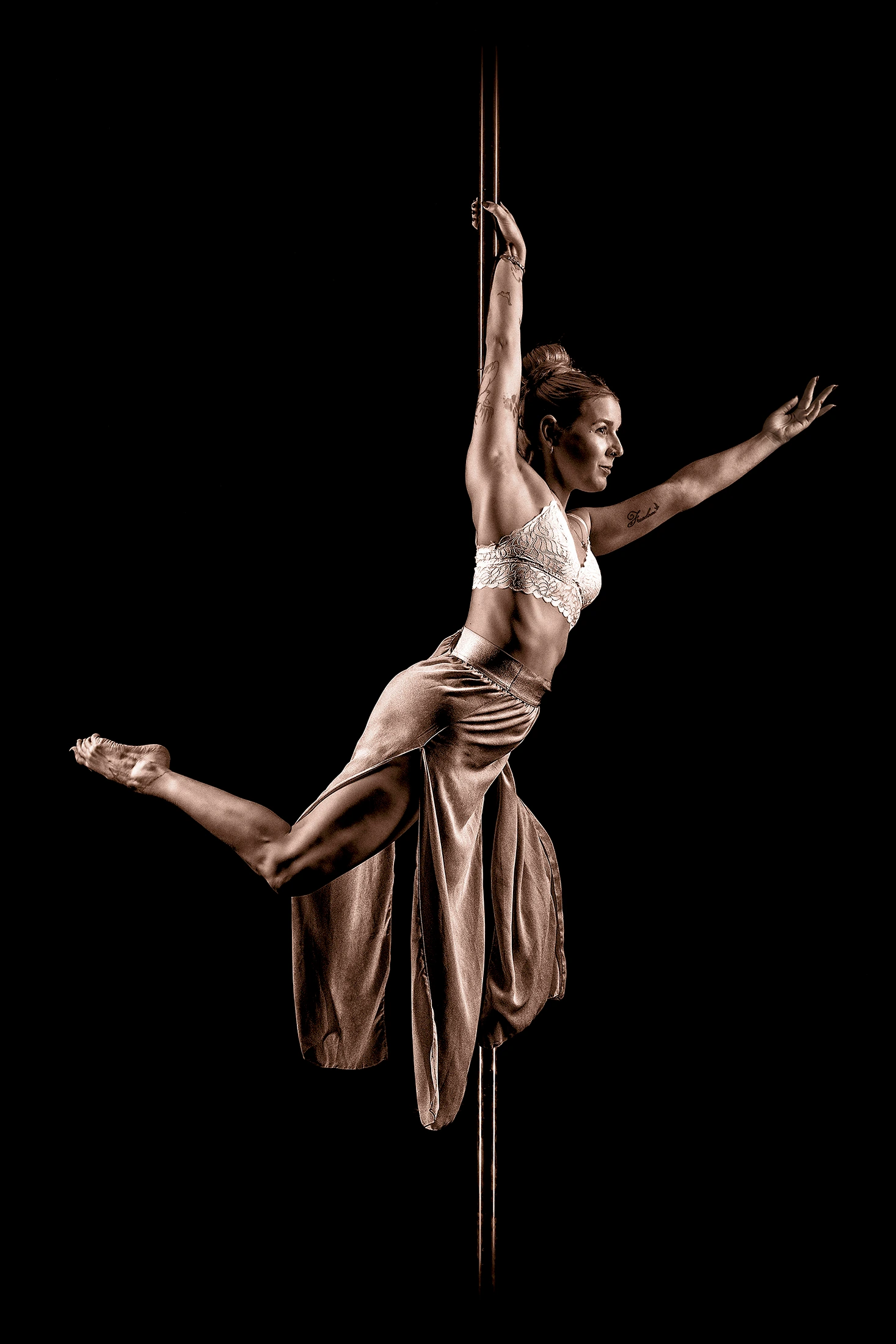 Pole artist hanging from one hand high on the pole, the other arm stretched wide, one leg reaching forward and loose wide trousers flowing below.